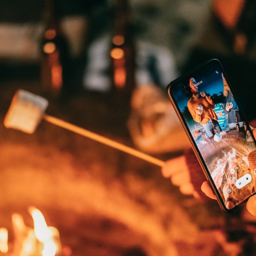 person takes photo on smart phone of group roasting marshmallows over fire 