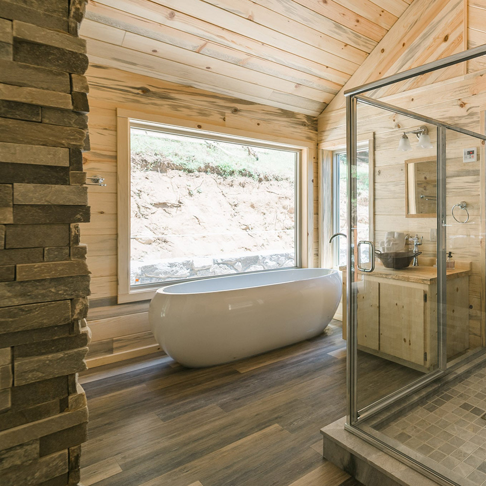 Rustic wood-paneled bathroom featuring a large freestanding white soaking tub in front of a picture window, a glass-enclosed shower, and a stone accent wall.